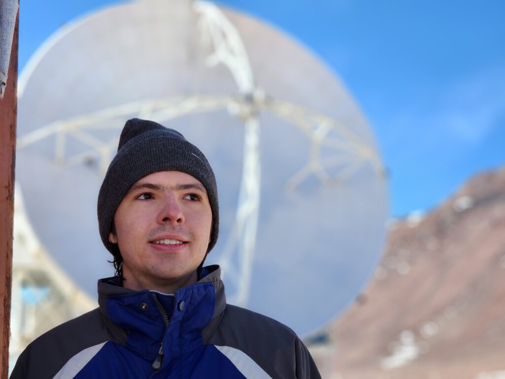 A photograph of Christopher Rooney with the APEX telescope in the background